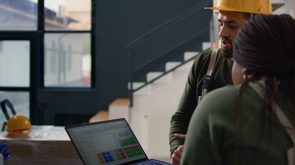 A man and woman in hard hats collaborate on a laptop, discussing plans using RFI Management Tools in a construction